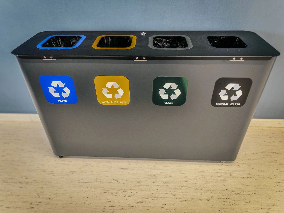 A four-section recycling bin with color-coded labels for paper, metal and plastic, glass, and general waste.
