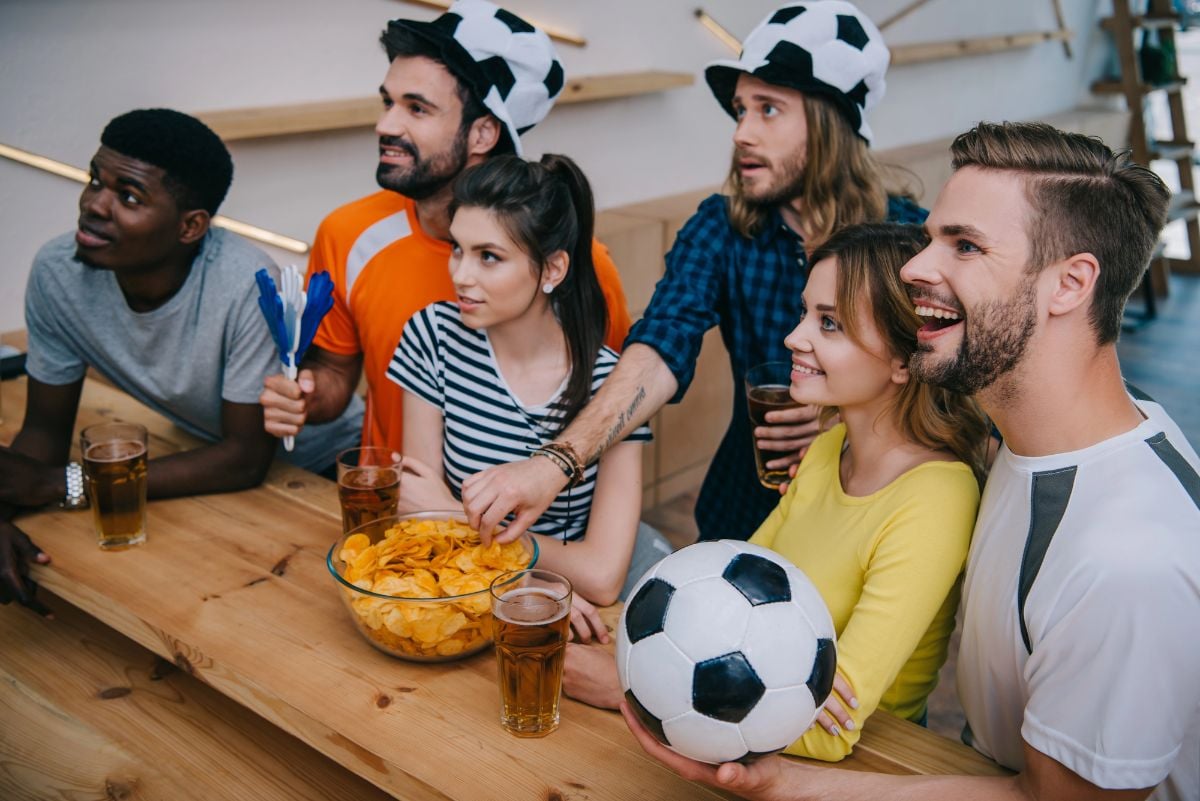 Group of friends in soccer ball hats drinking beer and watching football match.