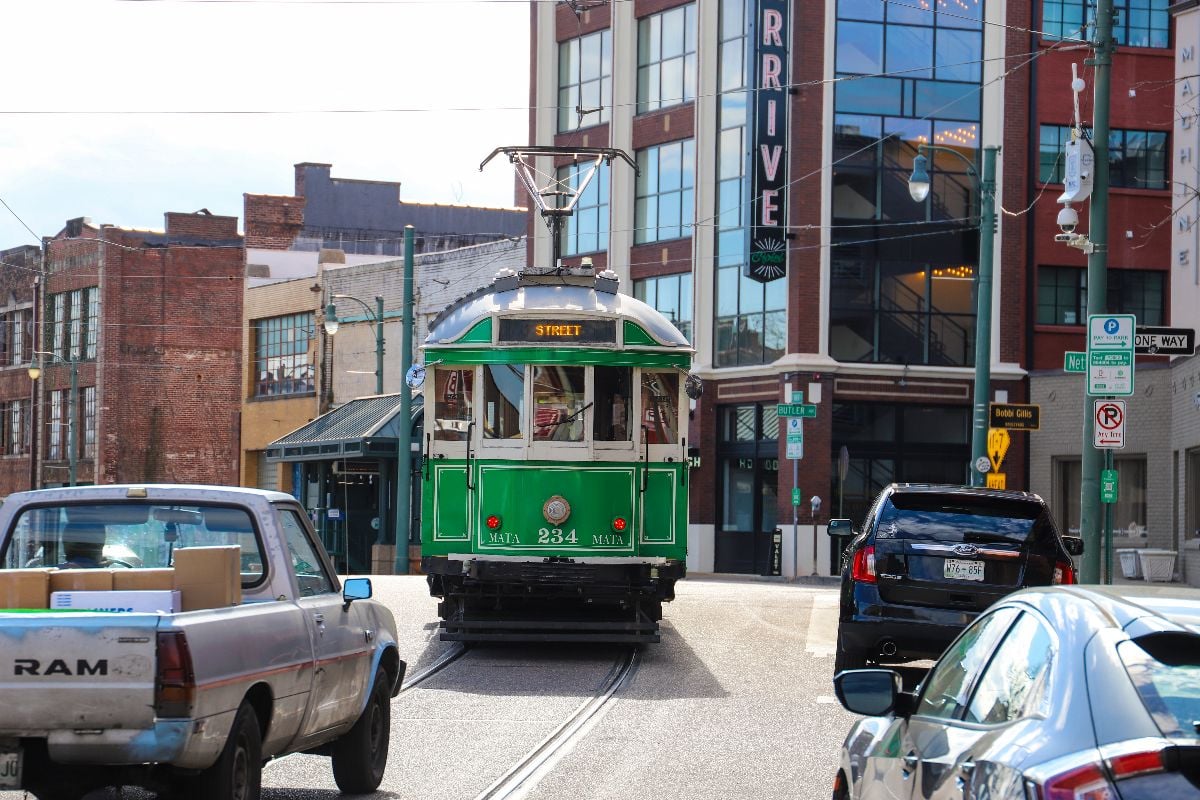 A green streetcars driving along a street with cars and trucks along the street.