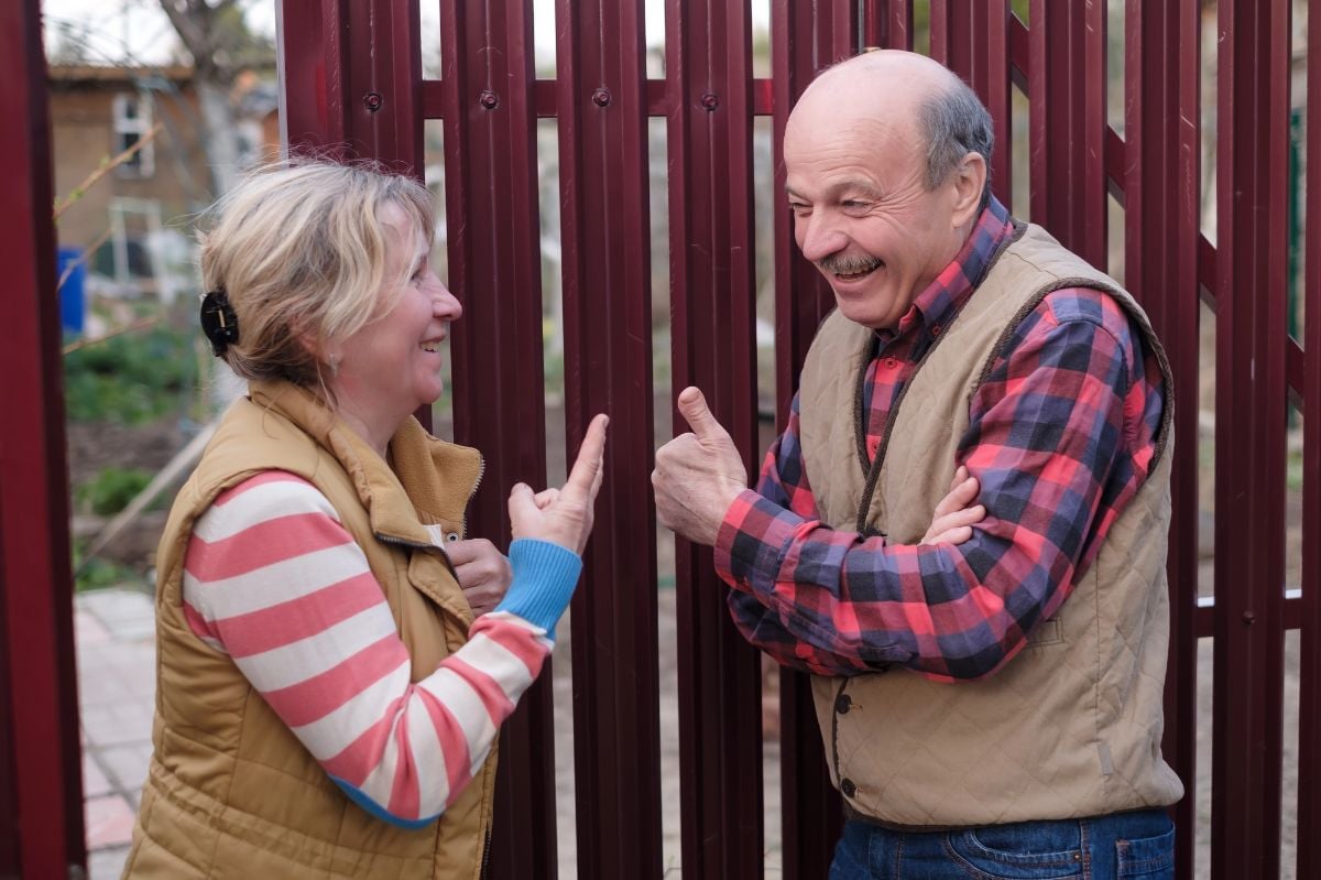 Two senior neighbors talking to each other on sunny day near fence.