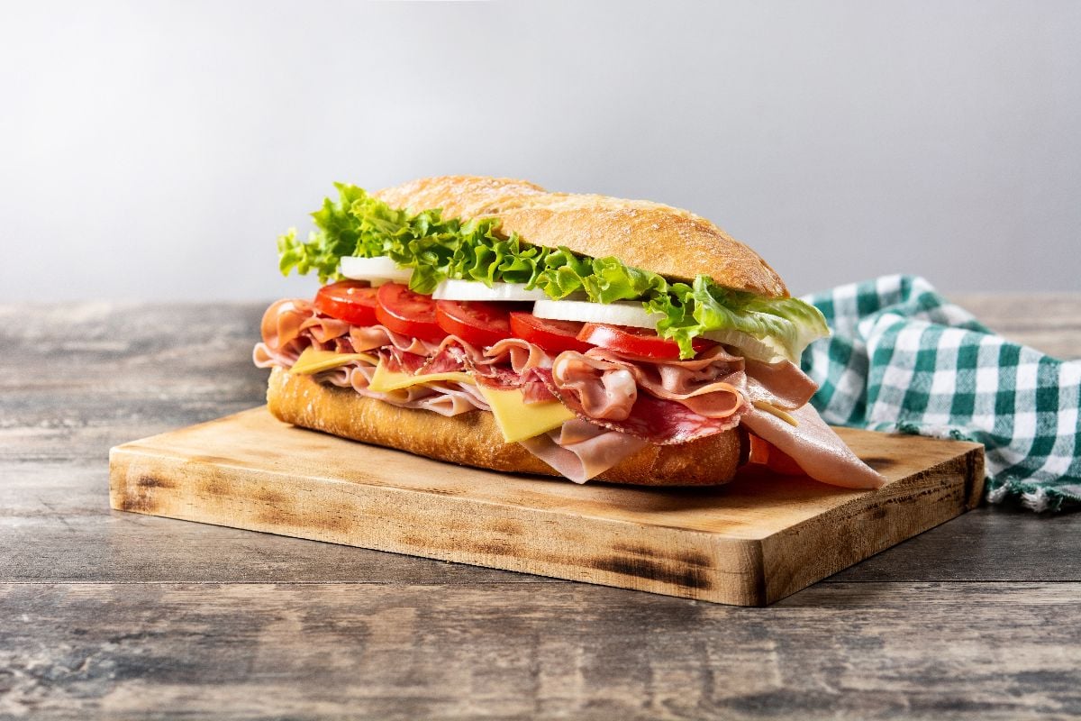 Submarine sandwich with ham, cheese, lettuce, tomatoes, onion, and sausage on wooden table.
