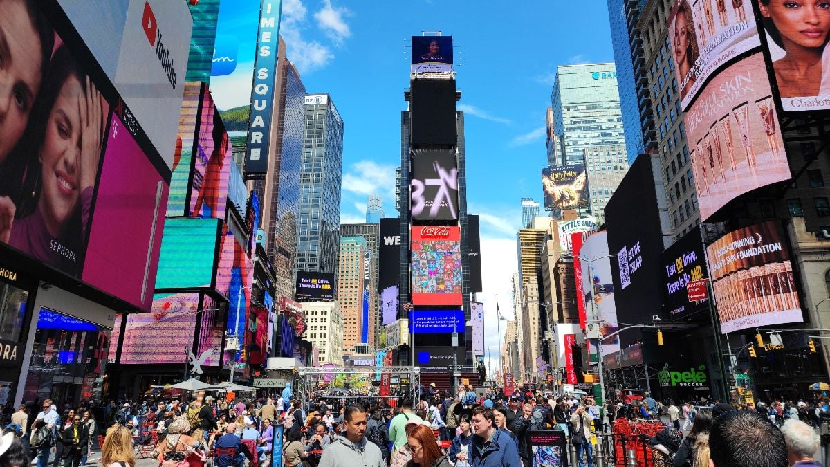 A large crowd of people fills Times Square, surrounded by towering buildings and bright digital billboards.