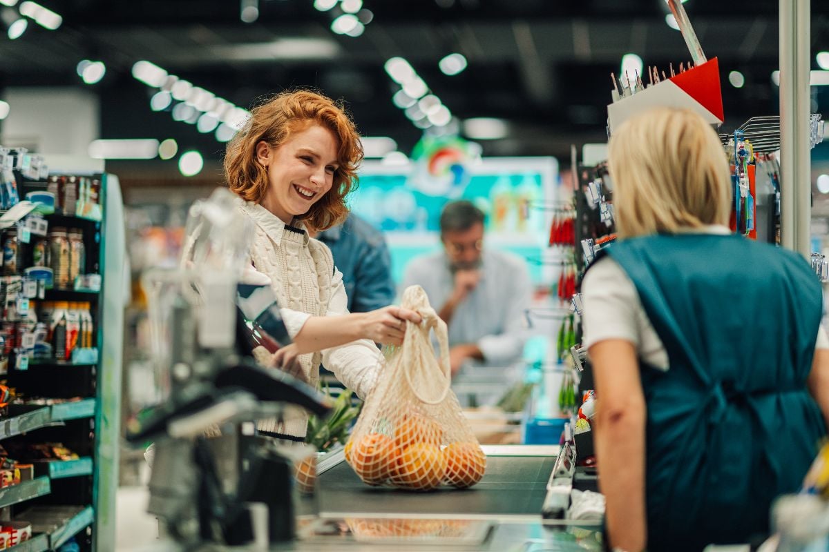 Customer placing reusable shopping bag with oranges on conveyor belt at supermarket checkout.