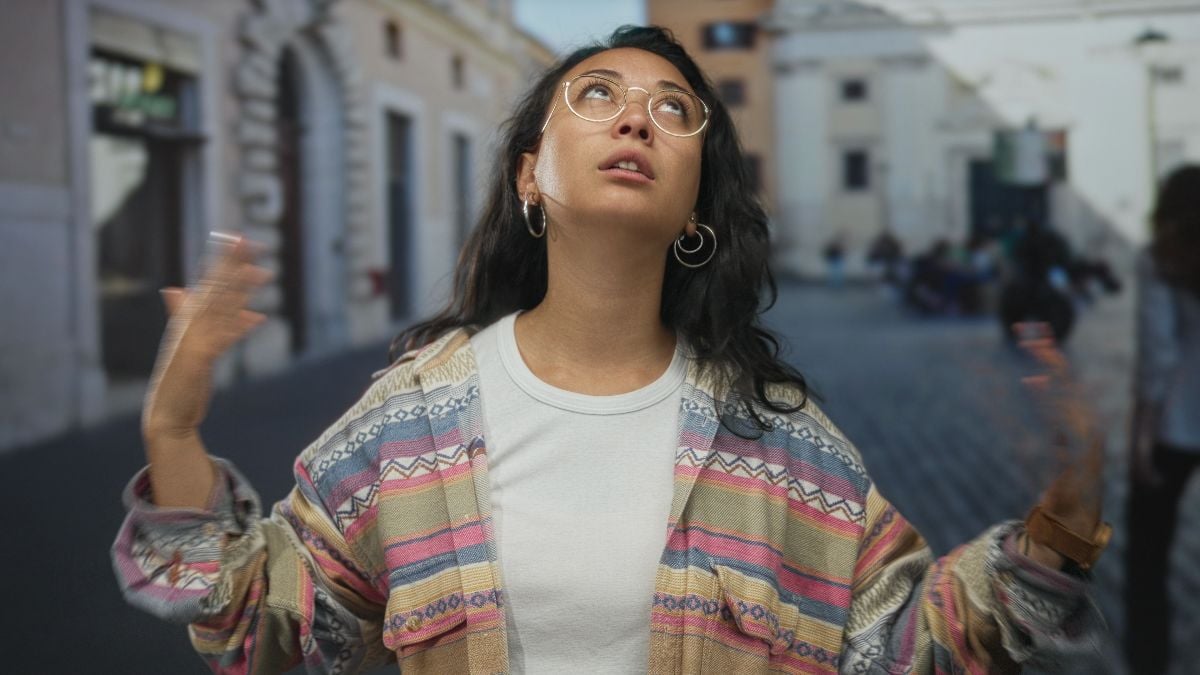 Woman touches hair with one hand and shrugs on a cobblestone street by buildings.