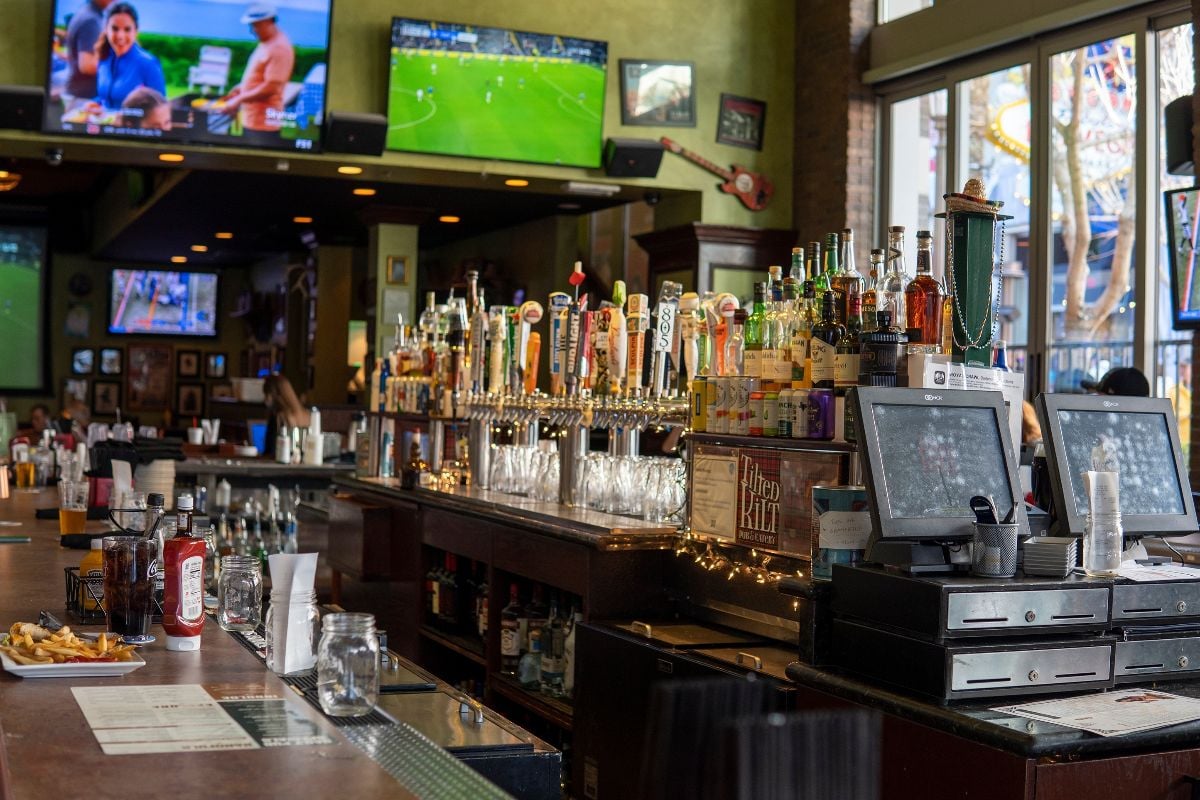 Bartender's place in a pub with shelves and bottles of alcohol and big screen televisions with sports.