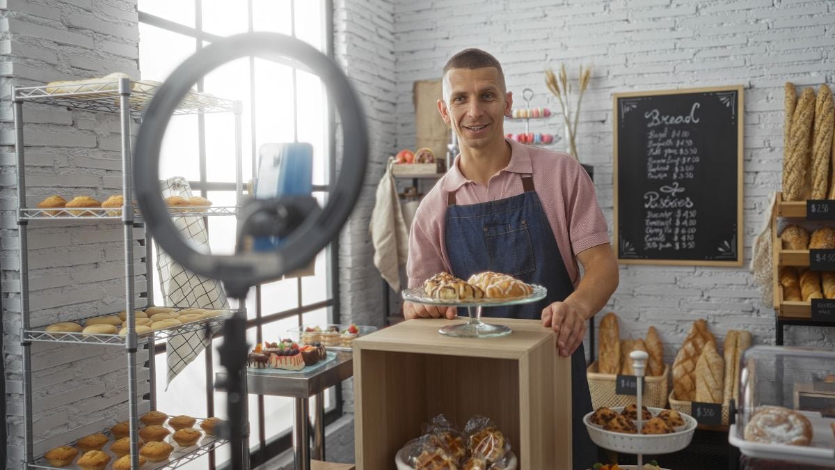 Man in bakery holding plate of pastries,, while posing in front of a filming setup with a ring light and camera.