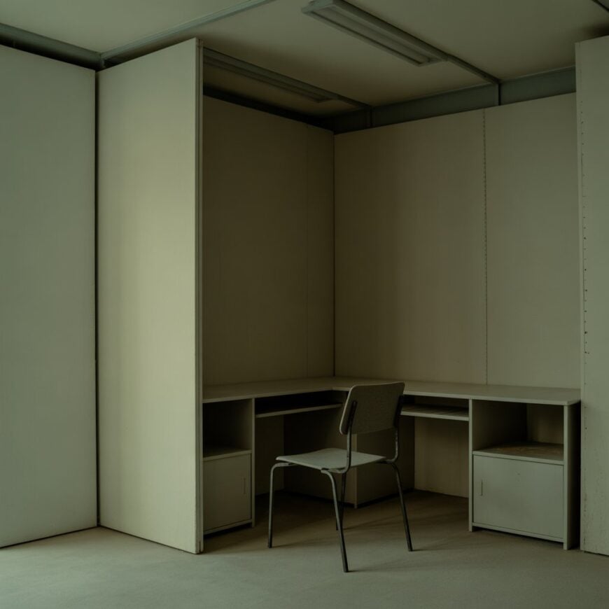 Empty beige cubicle with a simple desk and a single metal chair.