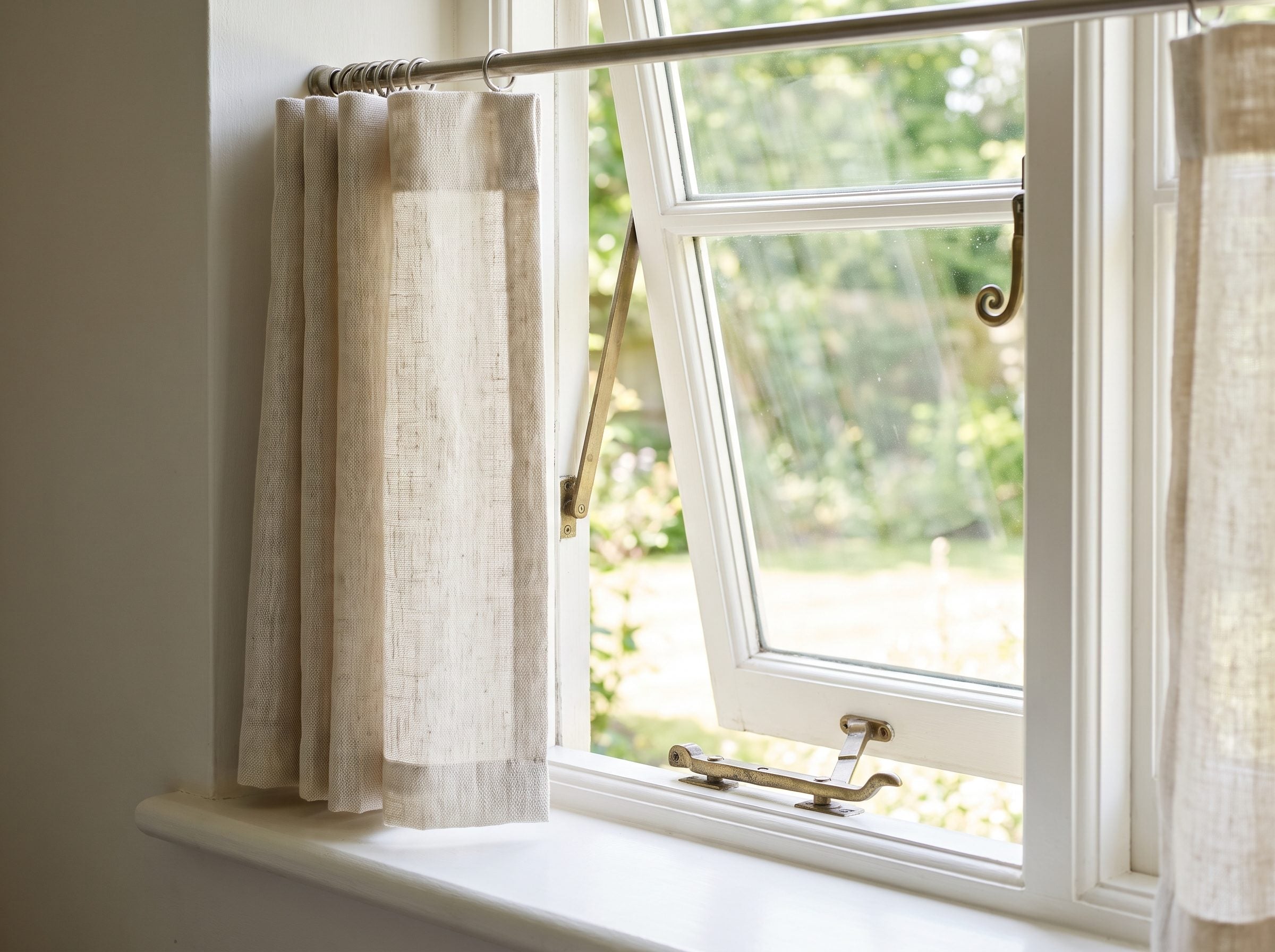 Kitchen casement window with sill-length linen panels stopping an inch above the sill and the crank handle left exposed