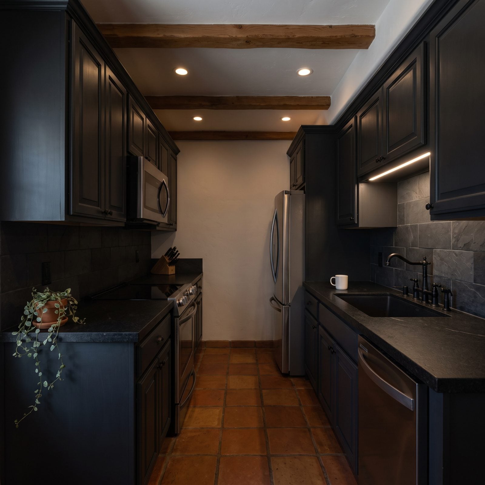 Windowless galley kitchen with charcoal cabinets and near-black granite counters that read dim under recessed lights.