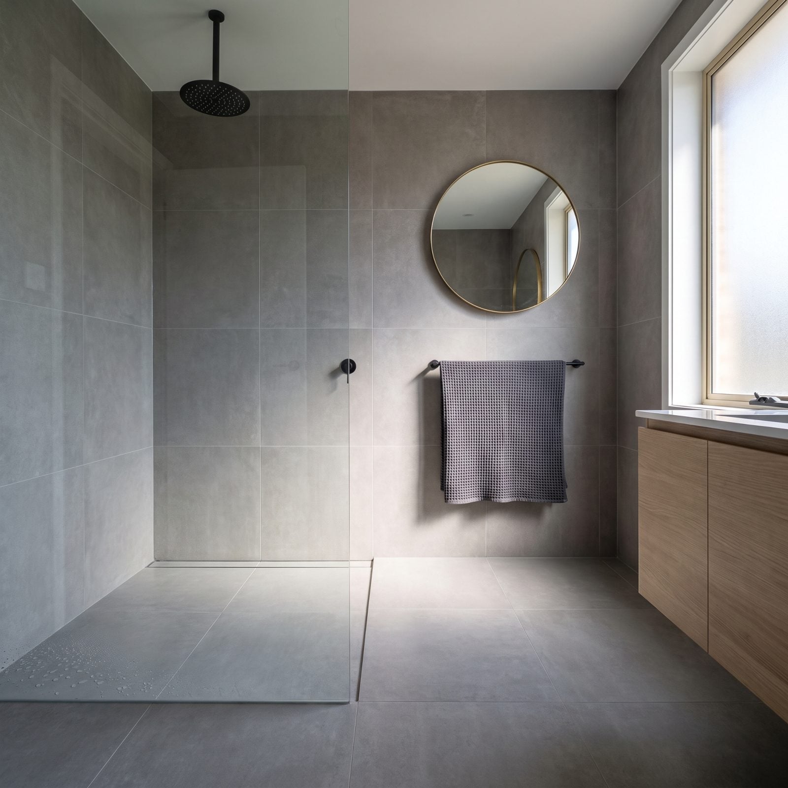 Gray-tiled curbless glass shower where water pools in a corner and moisture shadows the grout at the floor line.