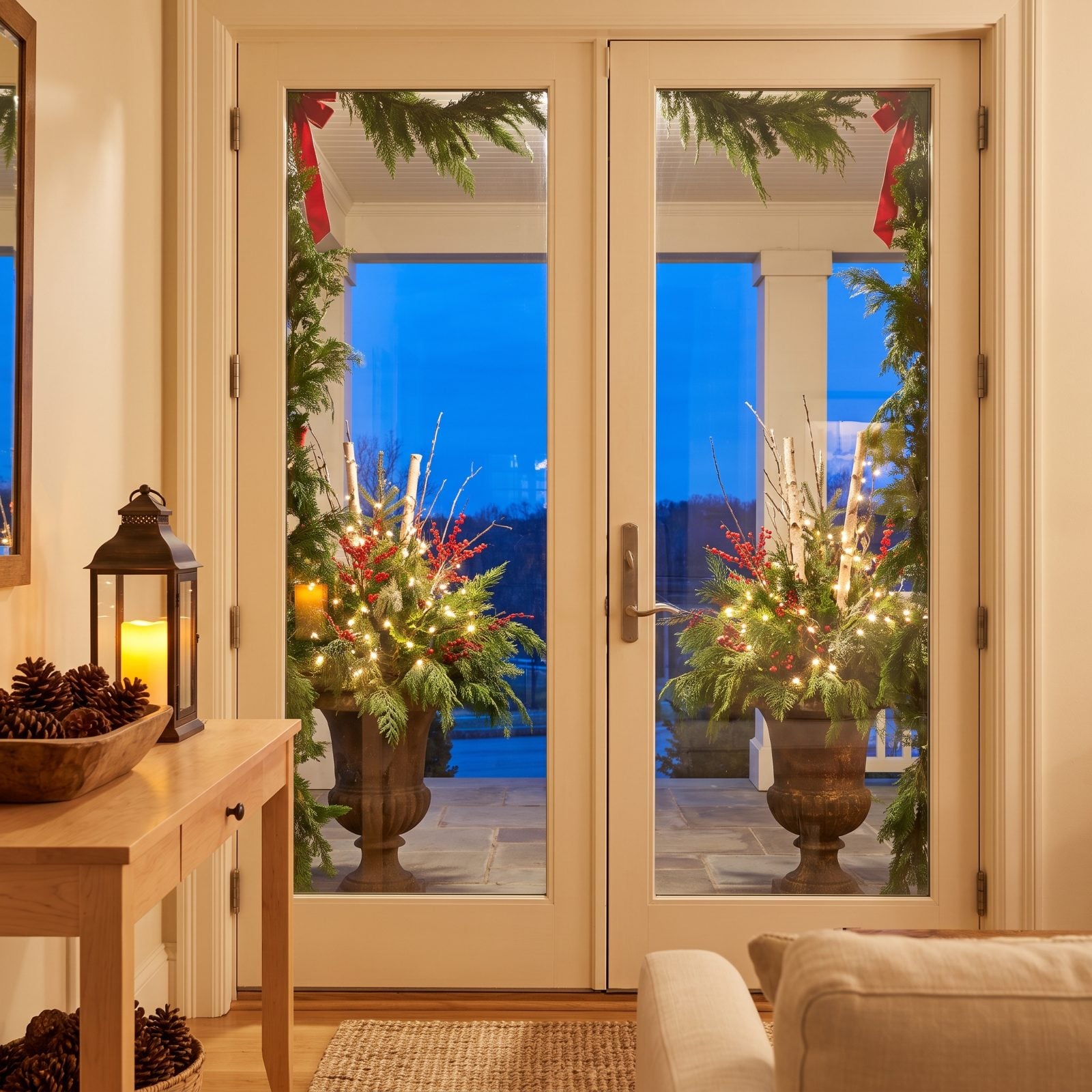 A Porch Visible Through French Doors With Lit Evergreen Urns