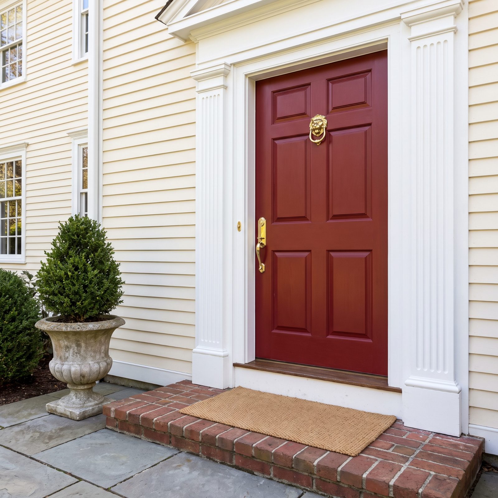 Traditional front entry with a deep red six-panel door, a brass knocker, and an evergreen in a stone urn to the left.