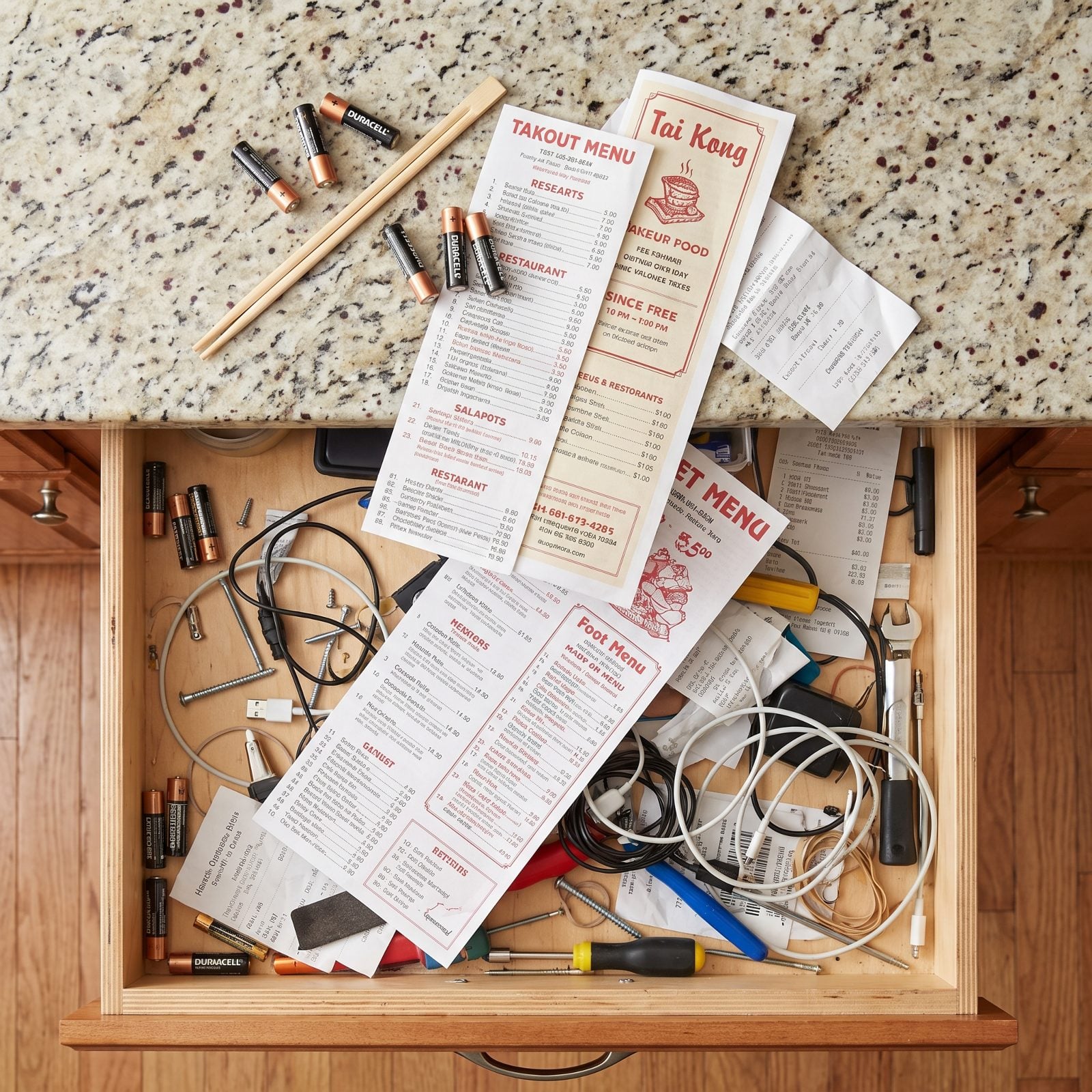 Kitchen with a half-open junk drawer holding batteries, rubber bands, and menus, a few items resting on the counter above it.